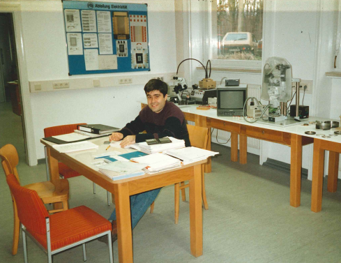 A young man at a simple table in front of laboratory equipment and a tube monitor.