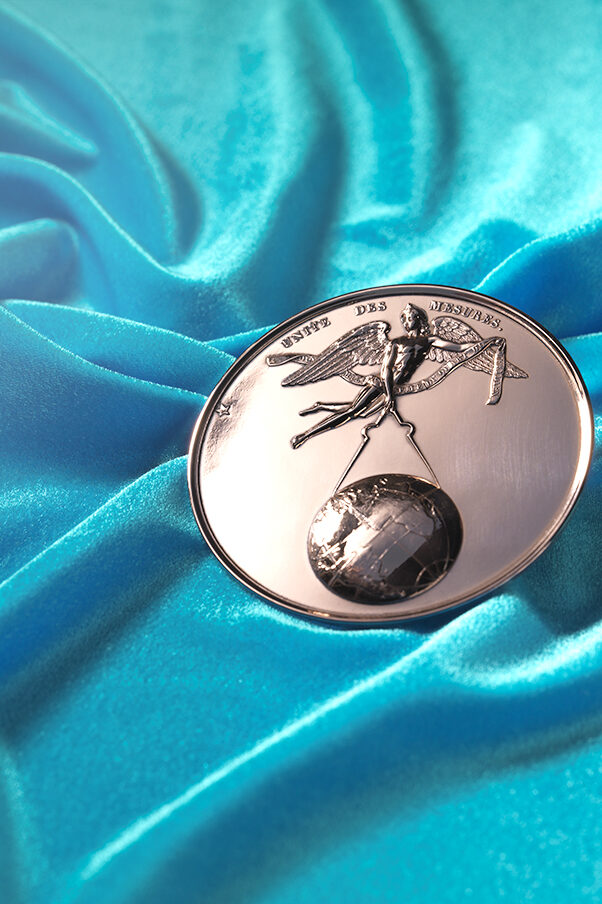 A medal made of copper showing a person with wings. This person is a genius holding a circle in their hand which is used to measure the world globe.