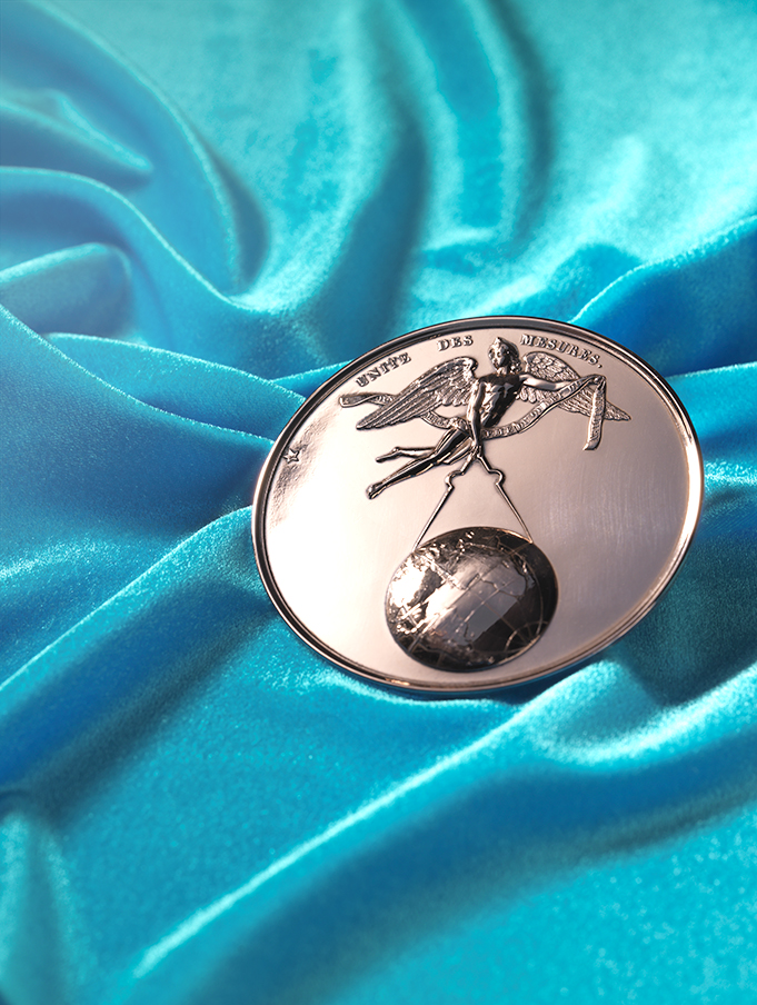 A medal made of copper showing a person with wings. This person is a genius holding a circle in their hand which is used to measure the world globe.