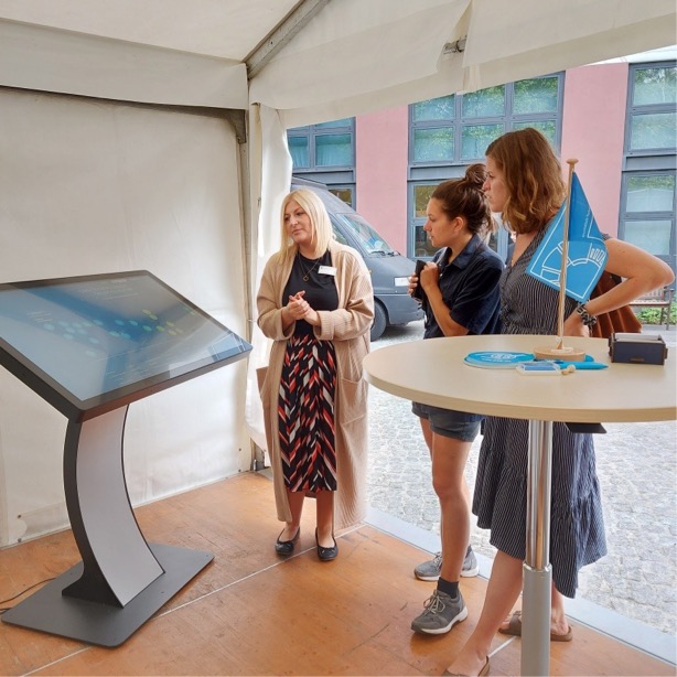 Three women stand in front of a touchscreen.