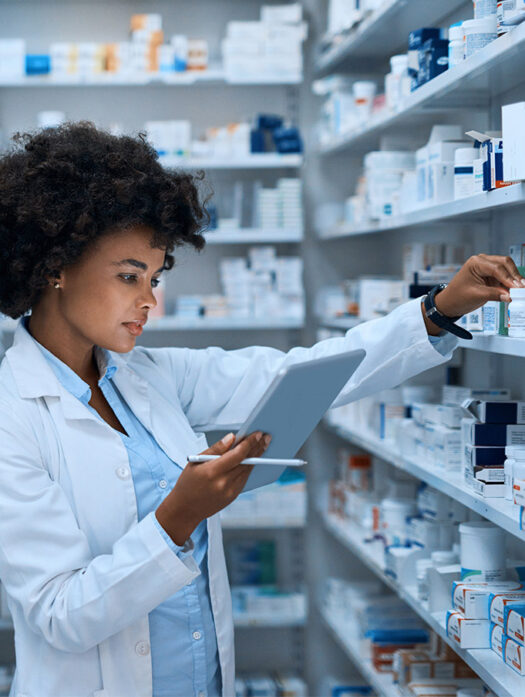 A woman in a white lab coat looks at the tablet in her hand. She is standing in front of a shelf with numerous medications.