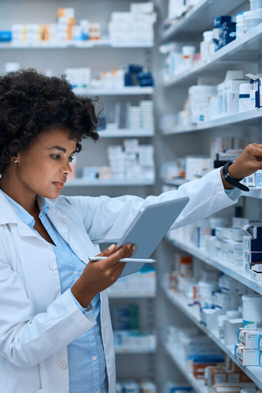 A woman in a white lab coat looks at the tablet in her hand. She is standing in front of a shelf with numerous medications.