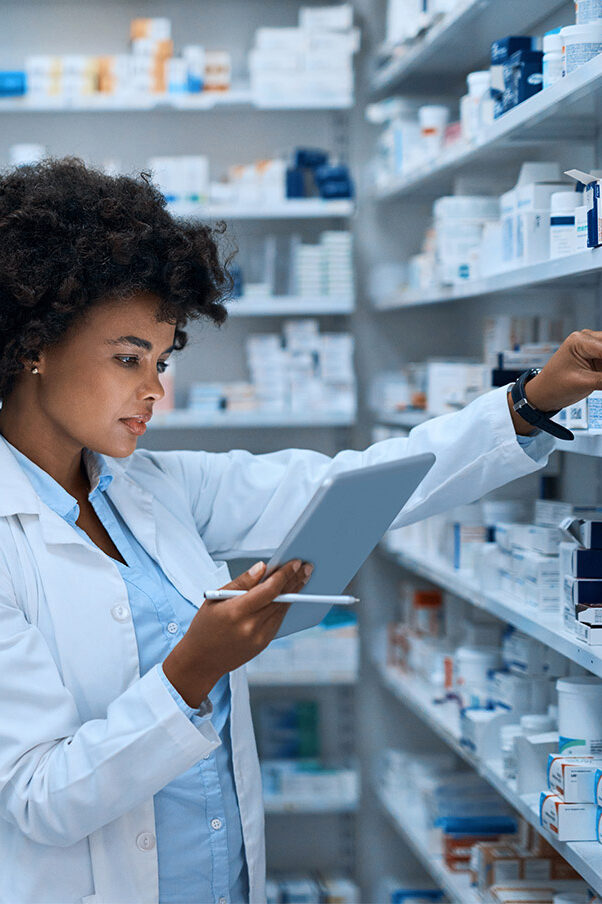 A woman in a white lab coat looks at the tablet in her hand. She is standing in front of a shelf with numerous medications.