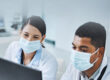 A man and a woman in white lab coats and wearing face masks sit in front of a laptop in a laboratory.