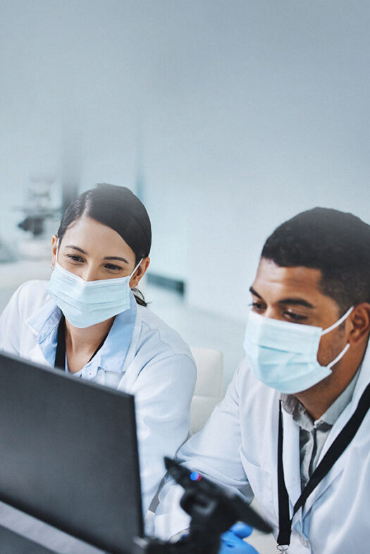 A man and a woman in white lab coats and wearing face masks sit in front of a laptop in a laboratory.