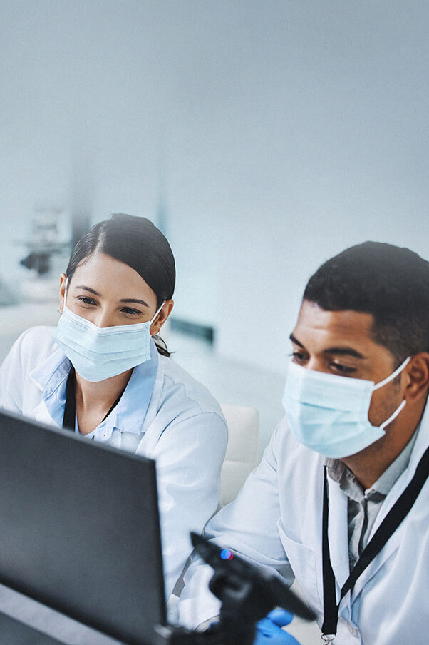 A man and a woman in white lab coats and wearing face masks sit in front of a laptop in a laboratory.