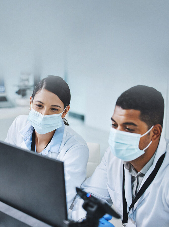 A man and a woman in white lab coats and wearing face masks sit in front of a laptop in a laboratory.