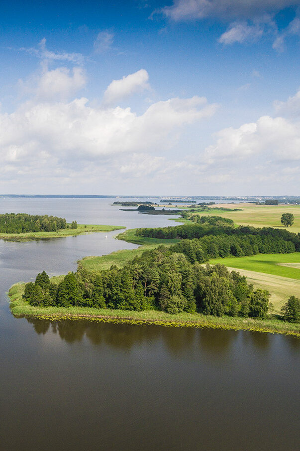 Photo of the Masurian Lake District in Poland. You can see a wide lake landscape with green shores and trees.