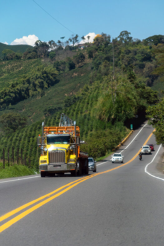 Un camión amarillo, cuatro coches y una moto circulan por una carretera rural en Colombia