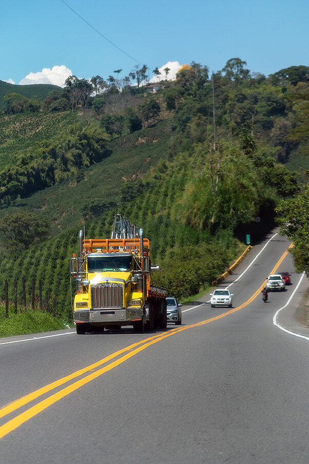 A yellow truck, four cars and a motorbike drive along a country road in Colombia