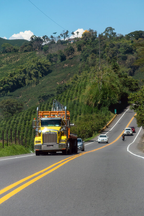 Un camión amarillo, cuatro coches y una moto circulan por una carretera rural en Colombia