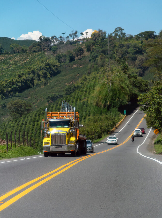 A yellow truck, four cars and a motorbike drive along a country road in Colombia