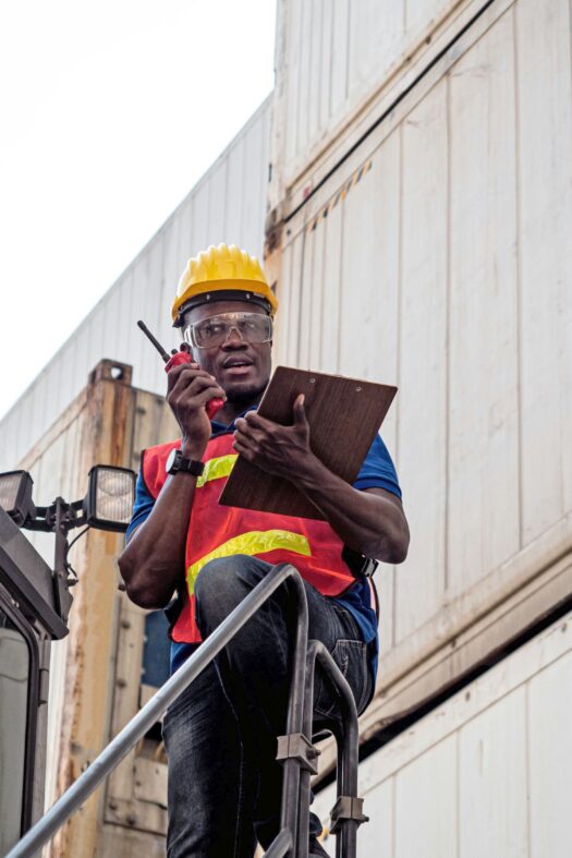 A foreman with yellow helmet and red waistcoat, holding a clipboard and speaking into a walkie-talkie.