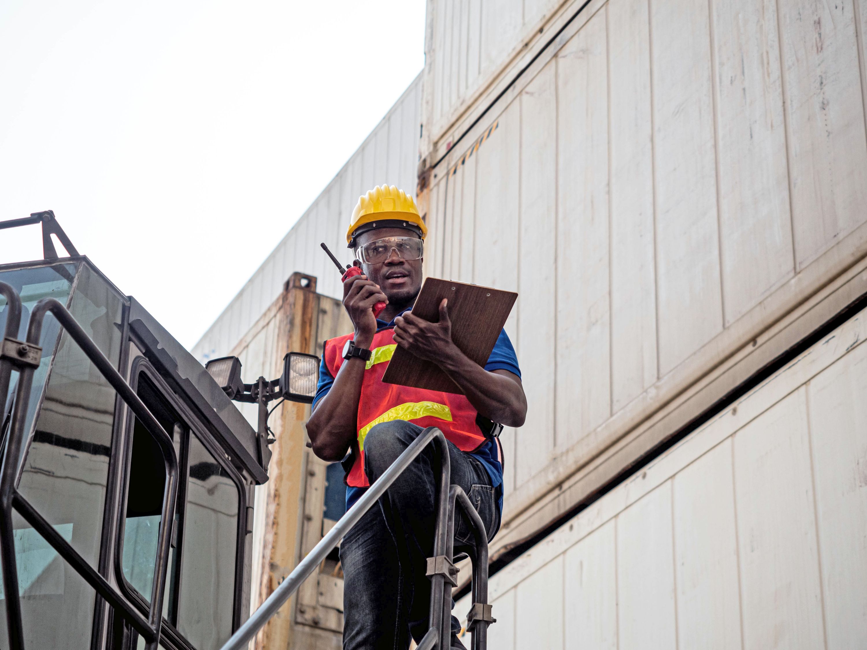 A foreman with yellow helmet and red waistcoat, holding a clipboard and speaking into a walkie-talkie.