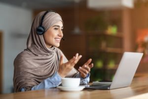 Young woman in hijab having video conference at cafe, using headset and laptop, drinking coffee, empty space