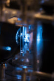 Foto einer Durchflussküvette in einem Laserdurchflusszytometer. Die Zellen strömen vertikal durch die Küvette und der Laser strahlt horizontal und ist in die Mitte des Durchflusskanals fokussiert. Links ist ein Teil des Objektivs zur Detektion zu sehen, das auf den Laserfokuspunkt ausgerichtet ist.