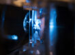Foto einer Durchflussküvette in einem Laserdurchflusszytometer. Die Zellen strömen vertikal durch die Küvette und der Laser strahlt horizontal und ist in die Mitte des Durchflusskanals fokussiert. Links ist ein Teil des Objektivs zur Detektion zu sehen, das auf den Laserfokuspunkt ausgerichtet ist.