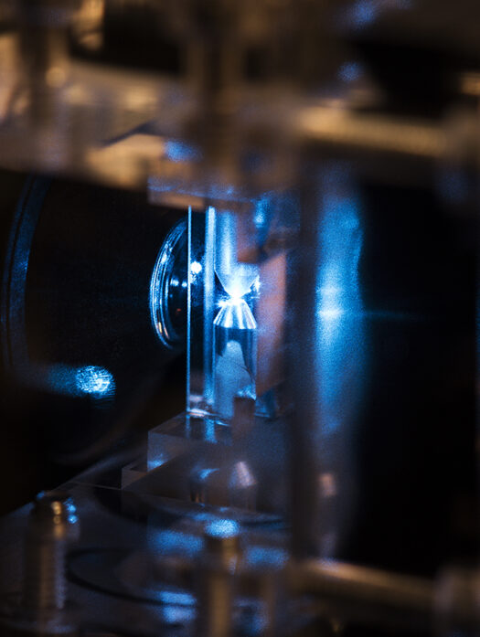 Photo of a flow-through cuvette in a laser flow cytometer. The cells flow vertically through the cuvette and the laser shines horizontally and is focused in the centre of the flow channel. On the left is part of the detection lens aligned with the laser focus point.