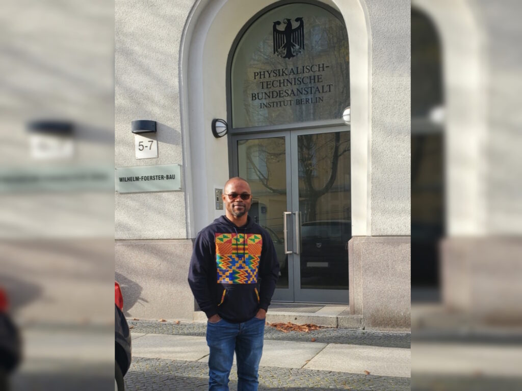 Mann mit buntem Pullover und Sonnenbrille steht vor einem Eingang eines Gebäudes, dessen Portal der Bundesadler samt der Aufschrift „Physikalisch-Technische Bundesanstalt Institut Berlin“ ziert.