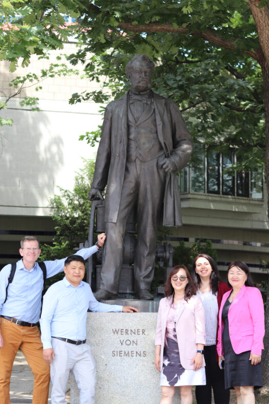 Two men and three women are standing next to a statue. The statue is of Werner von Siemens.