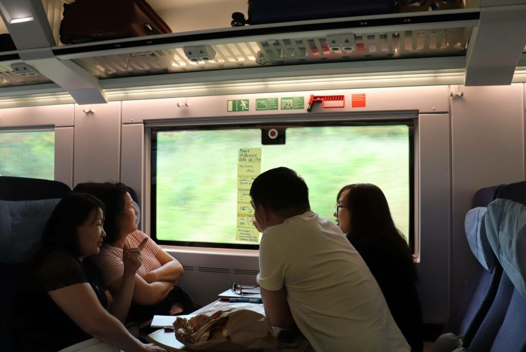 Four people sit across from each other on a train. They are looking at the window where a number of notes are taped.