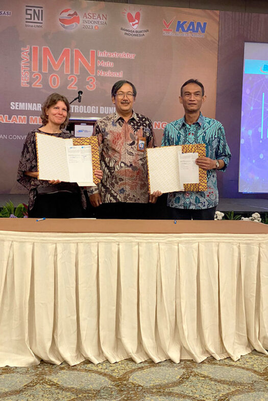 A woman and two men are standing behind a table. The woman and one man each hold an open folder with a document towards the camera.