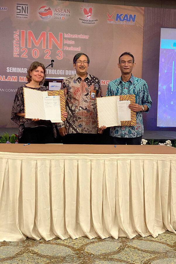 A woman and two men are standing behind a table. The woman and one man each hold an open folder with a document towards the camera.