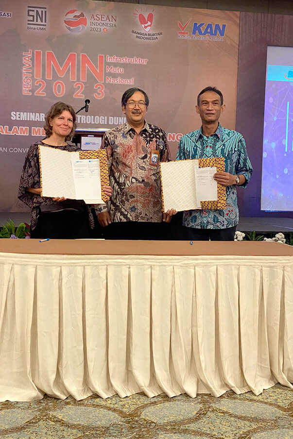 A woman and two men are standing behind a table. The woman and one man each hold an open folder with a document towards the camera.