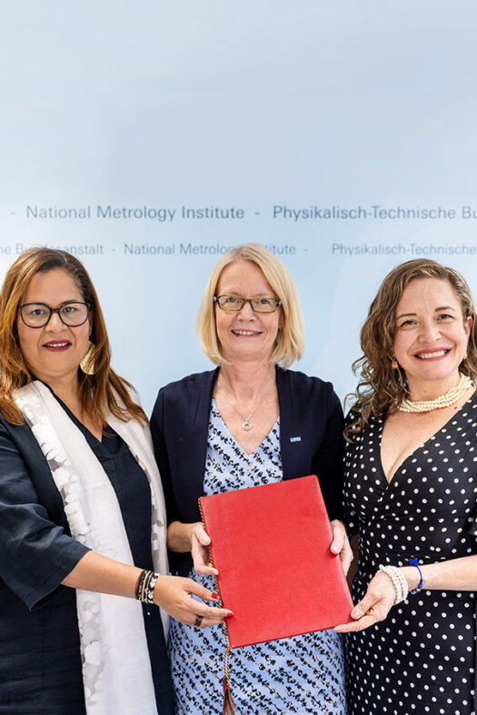 Three women stand next to each other. The woman in the centre holds a red folder in her hand and shows it to the camera. The women on the right and left touch the folder with one hand.