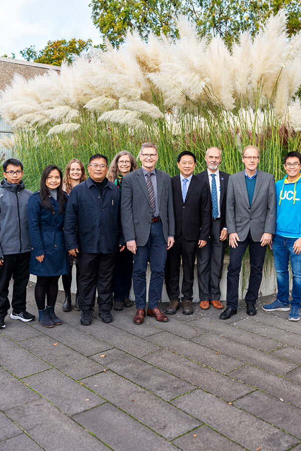 A group of men and women are standing outside in front of some green plants.