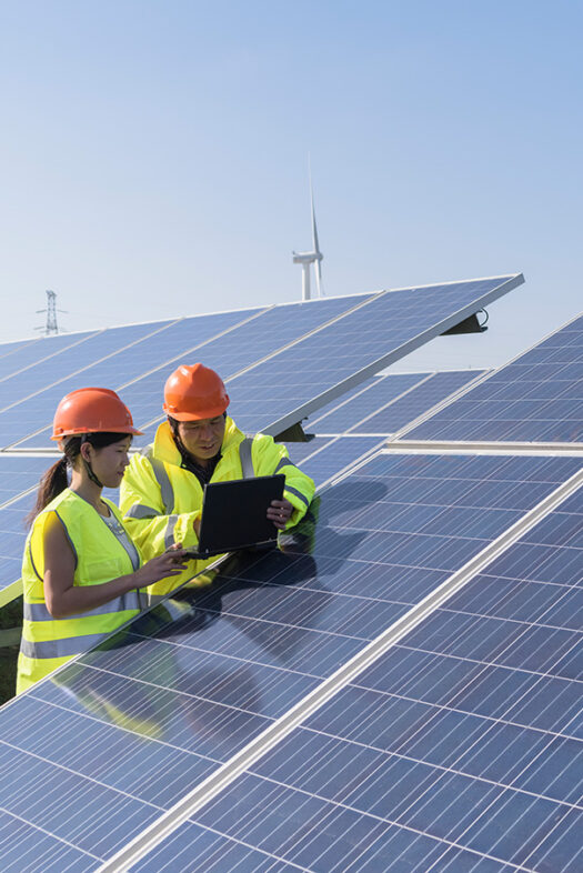 A woman and a man wearing safety helmets and yellow high-visibility waistcoats are standing in front of a photovoltaic system with a laptop.