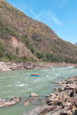 Der Oberlauf des Ganges. Links ist ein Hügelufer zu sehen, auf der rechten Seite ein kleiner Ausschnitt eines Felsenufers. Auf dem Fluss befindet sich ein kleines blaues Boot mit Menschen.