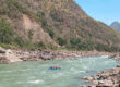 The upper reaches of the Ganges. On the left, a hilly riverbank can be seen; on the right, there is a rocky riverbank. A small blue boat with people inside is travelling down the river.