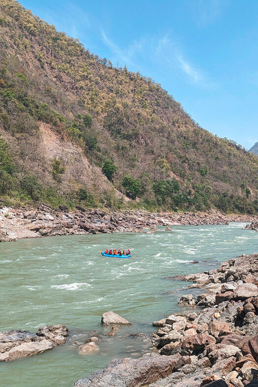 Der Oberlauf des Ganges. Links ist ein Hügelufer zu sehen, auf der rechten Seite ein kleiner Ausschnitt eines Felsenufers. Auf dem Fluss befindet sich ein kleines blaues Boot mit Menschen.