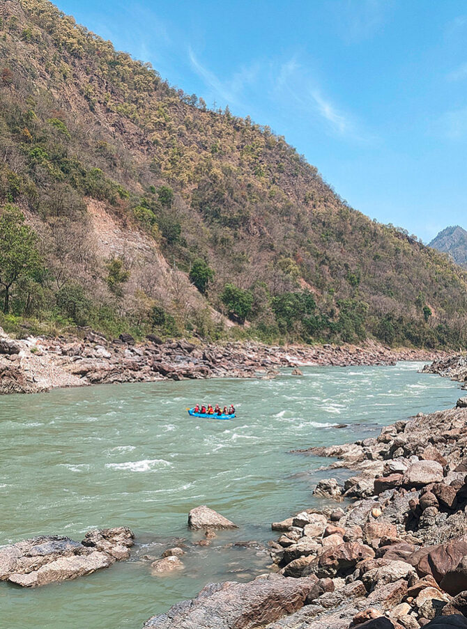 Der Oberlauf des Ganges. Links ist ein Hügelufer zu sehen, auf der rechten Seite ein kleiner Ausschnitt eines Felsenufers. Auf dem Fluss befindet sich ein kleines blaues Boot mit Menschen.