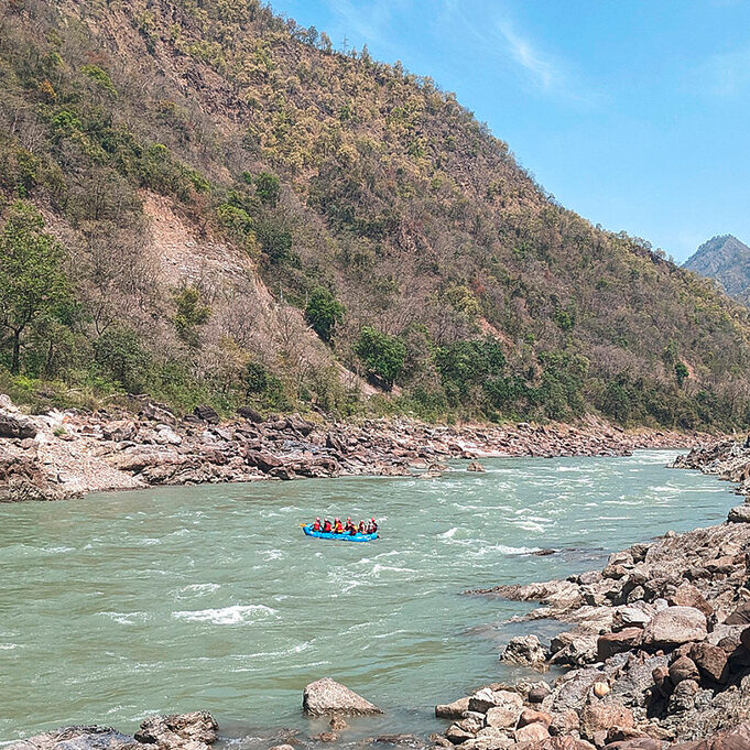 The upper reaches of the Ganges. On the left, a hilly riverbank can be seen; on the right, there is a rocky riverbank. A small blue boat with people inside is travelling down the river.