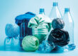 Vor einem blauen Hintergrund sind mehrere Kunststoffgegenstände zu sehen. Drei stehende Plastikflaschen, davor abgeschnittene Plastikflaschen, in denen zusammengerollte Stoffe in blau und grün stecken. Weiter vorne sind drei Wollknäule zu sehen und eine zusammengedrückte Plastikflasche.