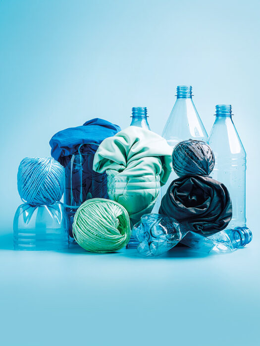Vor einem blauen Hintergrund sind mehrere Kunststoffgegenstände zu sehen. Drei stehende Plastikflaschen, davor abgeschnittene Plastikflaschen, in denen zusammengerollte Stoffe in blau und grün stecken. Weiter vorne sind drei Wollknäule zu sehen und eine zusammengedrückte Plastikflasche.