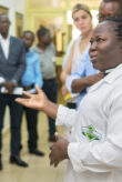 Eine Wissenschaftlerin, die einen Laborkittel der Ghana Standards Authority trägt, steht gestikulierend in einer Gruppe von Personen.