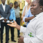 Une scientifique, portant une blouse de laboratoire de la Ghana Standards Authority, se tient debout, gesticulant, au milieu d'un groupe de personnes.