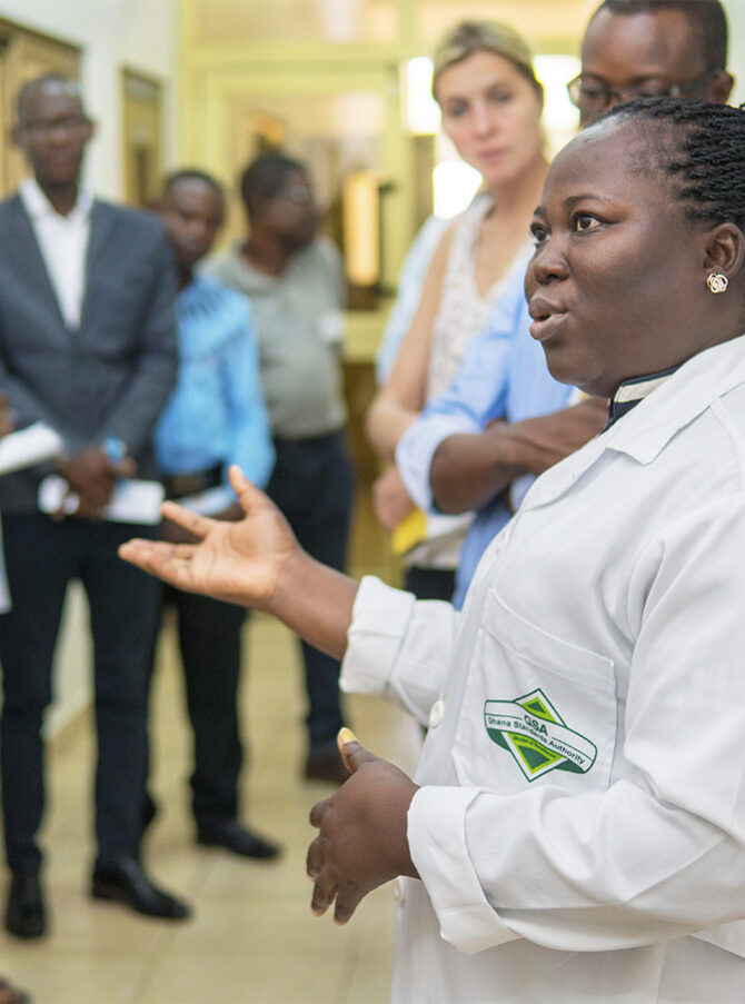 Une scientifique, portant une blouse de laboratoire de la Ghana Standards Authority, se tient debout, gesticulant, au milieu d'un groupe de personnes.