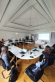Ein Konferenzsaal. An den Tischen, die in u-förmig angelegt sind, sitzen Personen. An der hinteren Wand wird eine Präsentation projiziert.
