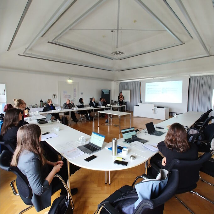 Ein Konferenzsaal. An den Tischen, die in u-förmig angelegt sind, sitzen Personen. An der hinteren Wand wird eine Präsentation projiziert.