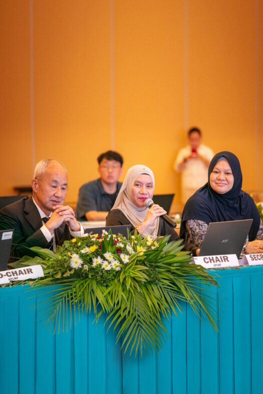 Two women wearing hijabs and a man are sitting at a table with flowers and a laptop. The woman in the middle is holding a microphone in her hand.