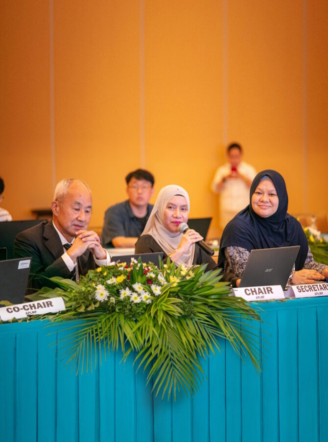 Two women wearing hijabs and a man are sitting at a table with flowers and a laptop. The woman in the middle is holding a microphone in her hand.
