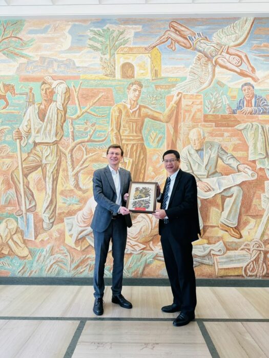 Two men are standing in front of a mural and holding up a framed painting together.