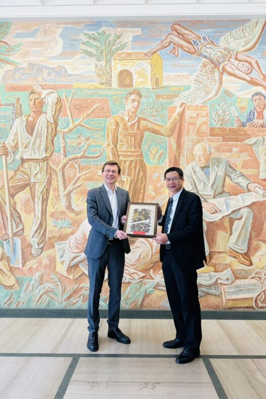 Two men are standing in front of a mural and holding up a framed painting together.