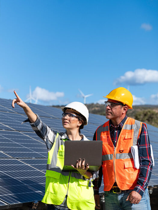 A man and a woman wearing safety helmets and high-visibility waistcoats stand in front of solar panels. The woman has a laptop in her hand and is pointing into the distance with her other hand.