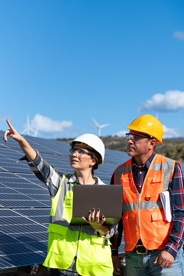 A man and a woman wearing safety helmets and high-visibility waistcoats stand in front of solar panels. The woman has a laptop in her hand and is pointing into the distance with her other hand.