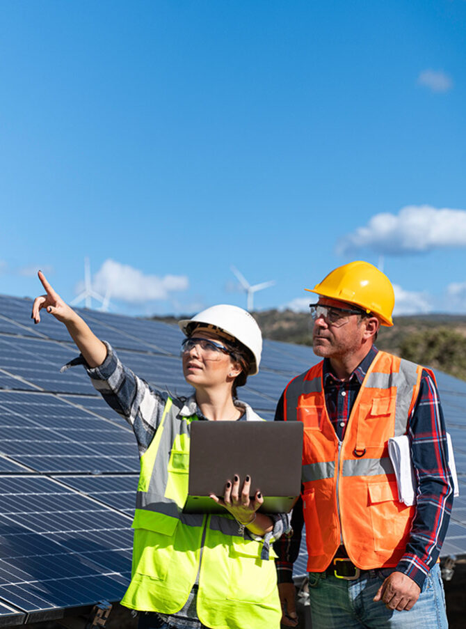 A man and a woman wearing safety helmets and high-visibility waistcoats stand in front of solar panels. The woman has a laptop in her hand and is pointing into the distance with her other hand.